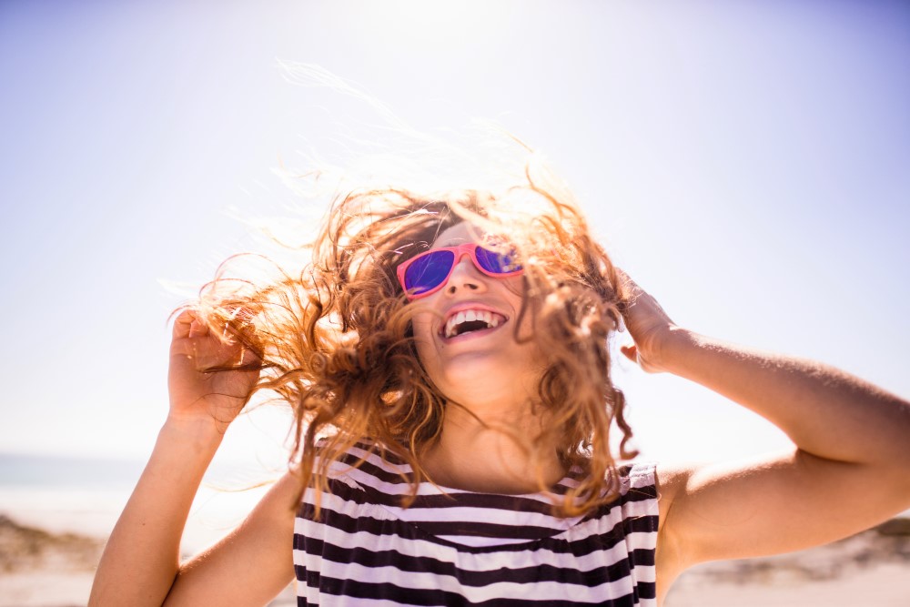 Eine glückliche Frau mit Sonnenbrille, im Hintergrund ist Strand zu sehen